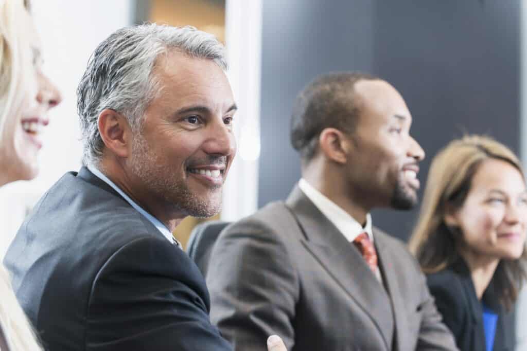 Close up of a mature businessman sitting with a group of multi-ethnic business people in a meeting. They are sitting in a row, watching something.