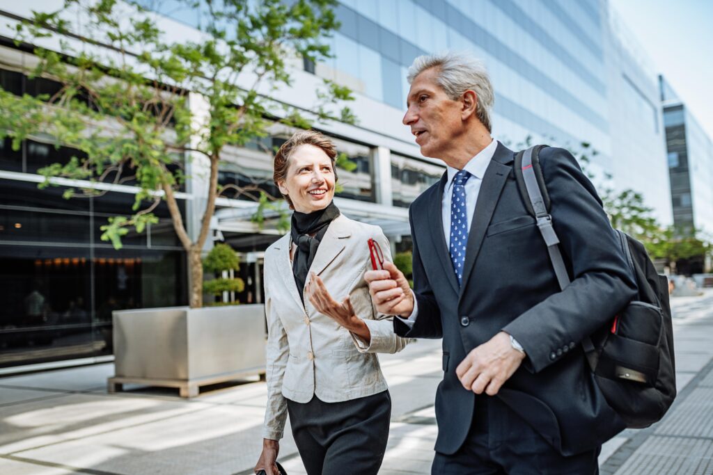 Two colleagues in suits walking outside side by side, passing an office and tree in the background 
