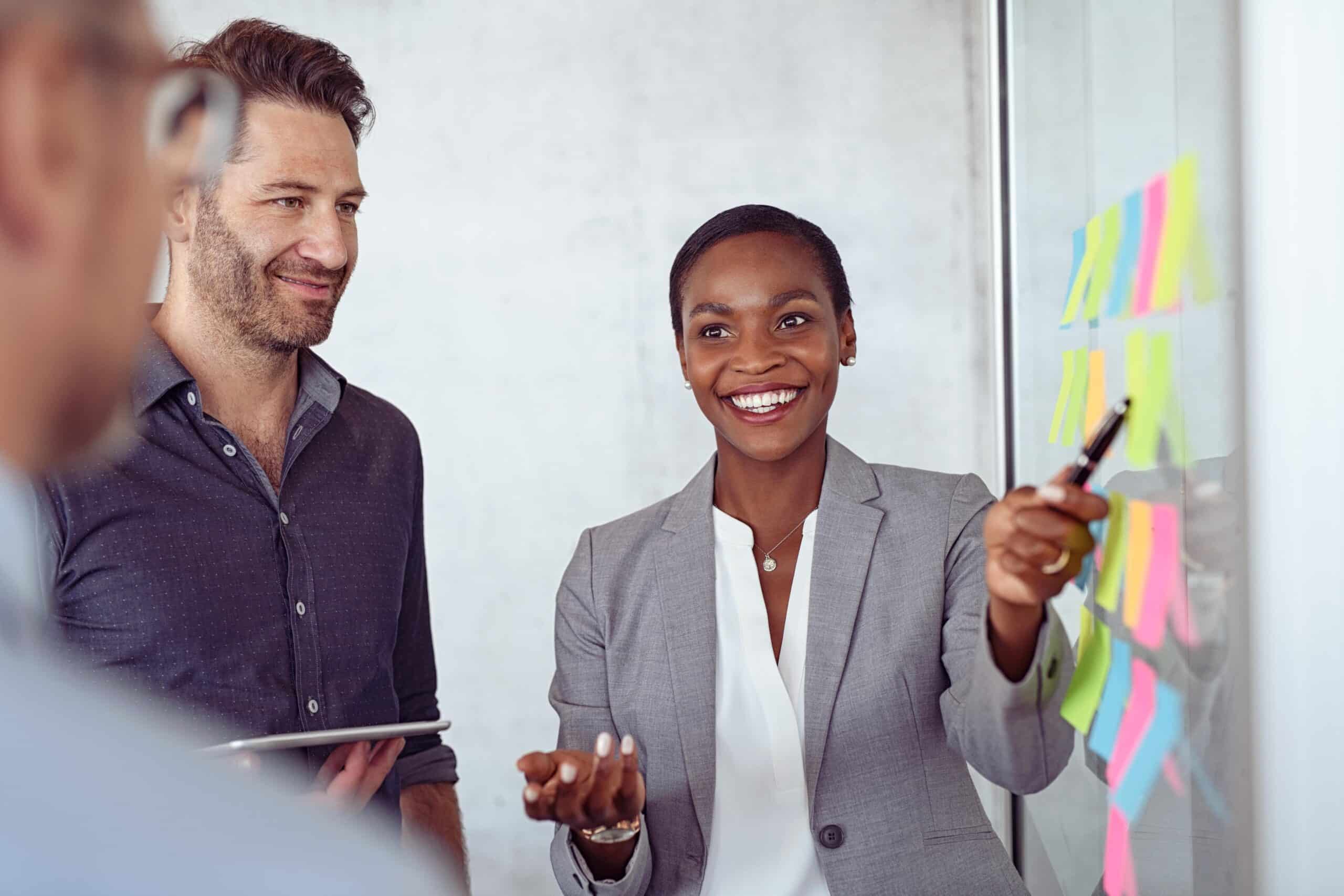 Smiling black businesswoman showing colorful sticky notes to business team on glass wall in modern office. Business people planning and discussing using post it notes in meeting room. Woman pointing with pen while explaining business plan to businessmen.