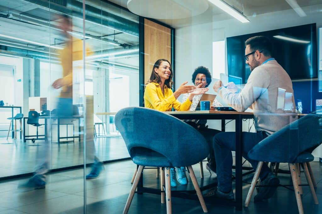 Businesspeople in Conference Room and Colleagues Walking By. Blurred motion of colleagues walking briskly down office hallway as colleagues sit in conference room discussing ideas. Busy modern workplace. Working process in the office, business people working, walking and talking, blurred motion